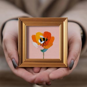 Puede incluir: Pequeña pintura enmarcada de una abeja en una flor. La flor es naranja, amarilla y blanca, con un tallo verde. La abeja es negra y blanca. El marco es de color dorado.