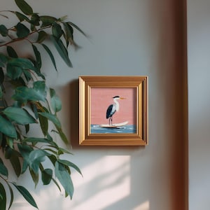 K&ouml;nnte beinhalten: Quadratisch gerahmtes Gem&auml;lde eines Reihers auf einem Surfbrett. Das Kunstwerk zeigt einen rosa Himmel, blaues Wasser und einen goldenen Rahmen. Der Reiher ist in Schwarz, Wei&szlig; und Blau dargestellt. Das Gem&auml;lde ist an einer hellgrauen Wand mit gr&uuml;nen Bl&auml;ttern im Hintergrund angebracht.