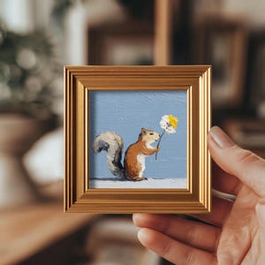 May include: A small framed painting of a squirrel holding a flower against a light blue backdrop. The squirrel is brown and white, and the flower has white and yellow petals. The frame is gold-coloured.