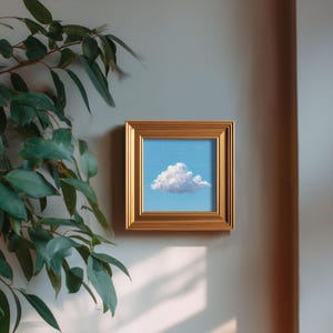 May include: Square framed painting of a fluffy white cloud against a light blue sky. The painting is in a gold-colored frame and is hanging on a light gray wall. Green leafy plant in the upper left corner.