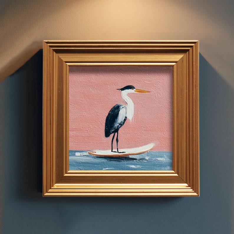 K&ouml;nnte beinhalten: Quadratisches gerahmtes Kunstwerk mit einem Reiher auf einem Surfbrett. Das Gem&auml;lde hat einen rosa Hintergrund, blaues Wasser und einen goldenen Rahmen. Der Reiher ist schwarz, wei&szlig; und grau mit einem orangefarbenen Schnabel.