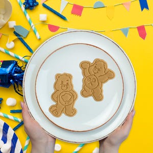 May include: Two brown bear-shaped biscuits on a white plate with a brown rim. The plate is held by hands against a yellow background with party decorations, including colourful flags and straws. The biscuits have detailed facial features and belly symbols.