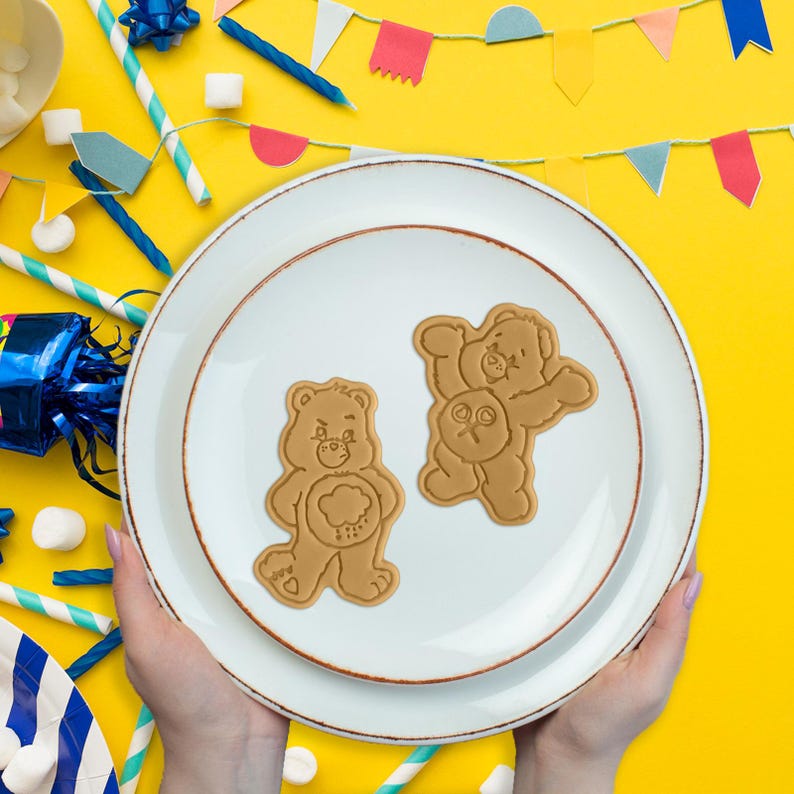 May include: Two brown bear-shaped biscuits on a white plate with a brown rim. The plate is held by hands against a yellow background with party decorations, including colourful flags and straws. The biscuits have detailed facial features and belly symbols.