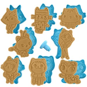 May include: A collection of tan and blue cat-shaped cookie cutters. The cutters feature various cat-themed designs, including a cat wearing a crown, a cat in a costume, and a cat with a mermaid tail. A small blue tool is also included.