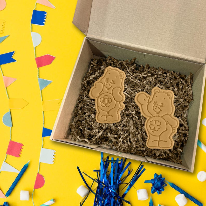 May include: Two tan Care Bear-shaped biscuits sit in a cardboard box filled with brown packing material. One biscuit has a flower on its belly, the other a four-leaf clover. The box is on a yellow surface with party decorations.