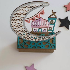 May include: A wooden decorative ornament featuring a crescent moon with a detailed geometric pattern, a mosque with a pink dome and minarets, and a teal base with a geometric pattern. The ornament is surrounded by stars.