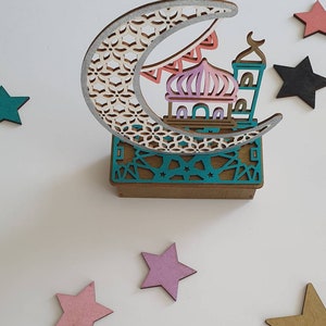 May include: A wooden decorative ornament featuring a crescent moon with a detailed geometric pattern, a mosque with a pink dome, and a teal base with a geometric pattern. The ornament is surrounded by five wooden stars in various colors.