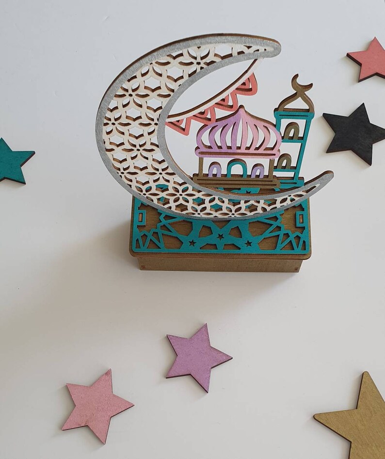 May include: A wooden decorative ornament featuring a crescent moon with a detailed geometric pattern, a mosque with a pink dome, and a teal base with a geometric pattern. The ornament is surrounded by five wooden stars in various colors.