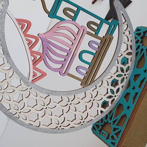 May include: A decorative wooden crescent moon with a white and silver patterned design. The moon is layered with a pink, teal, and brown mosque design. The mosque has a star and crescent moon on top.