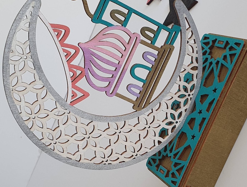 May include: A decorative wooden crescent moon with a white and silver patterned design. The moon is layered with a pink, teal, and brown mosque design. The mosque has a star and crescent moon on top.