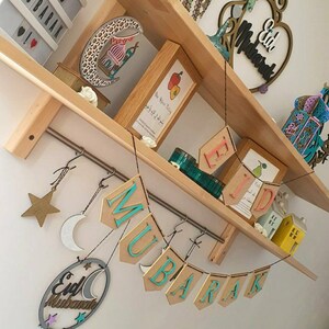 May include: A wooden shelf with a variety of decorations, including a crescent moon, a star, and a banner that reads "Eid Mubarak". The shelf is decorated with a variety of other items, including a small wooden house, a lantern, and a book.