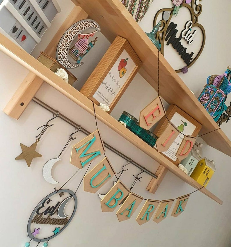 May include: A wooden shelf with a variety of decorations, including a crescent moon, a star, and a banner that reads "Eid Mubarak". The shelf is decorated with a variety of other items, including a small wooden house, a lantern, and a book.