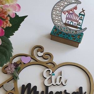 May include: A wooden heart-shaped sign with the words "Eid Mubarak" in black and white lettering. The sign is decorated with a gold frame, a pink heart, and a small green succulent. A wooden crescent moon with a mosque cutout is also pictured.