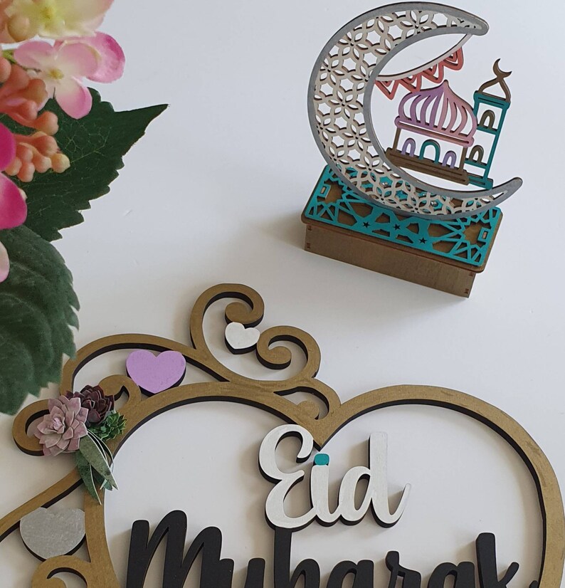 May include: A wooden heart-shaped sign with the words "Eid Mubarak" in black and white lettering. The sign is decorated with a gold frame, a pink heart, and a small green succulent. A wooden crescent moon with a mosque cutout is also pictured.