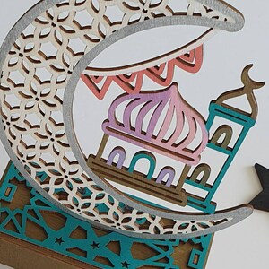 May include: A wooden decorative crescent moon with a detailed cut-out design, featuring a mosque with a pink and purple dome and a teal minaret. The moon is decorated with a silver trim and a string of colorful flags.