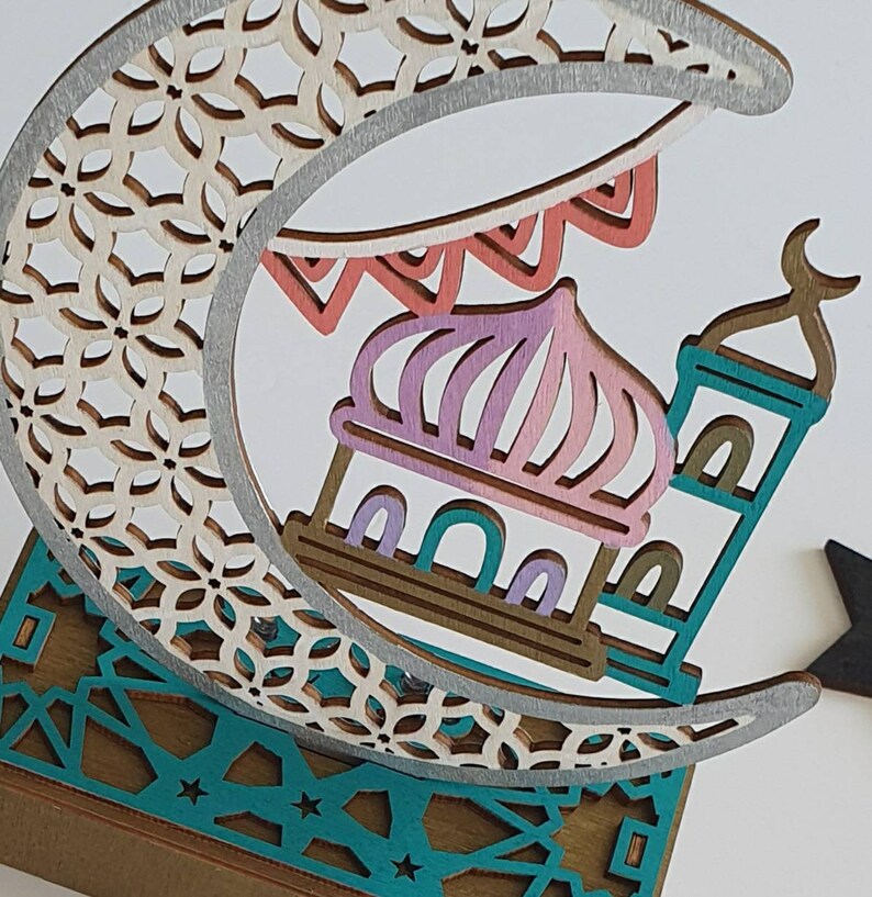 May include: A wooden decorative crescent moon with a detailed cut-out design, featuring a mosque with a pink and purple dome and a teal minaret. The moon is decorated with a silver trim and a string of colorful flags.
