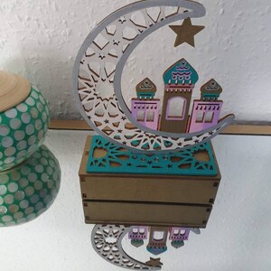 May include: A wooden crescent moon with a star and a small mosque-like structure inside. The moon is decorated with a white and silver geometric pattern. The mosque is painted in pink, teal, and brown. The piece is sitting on a wooden base.