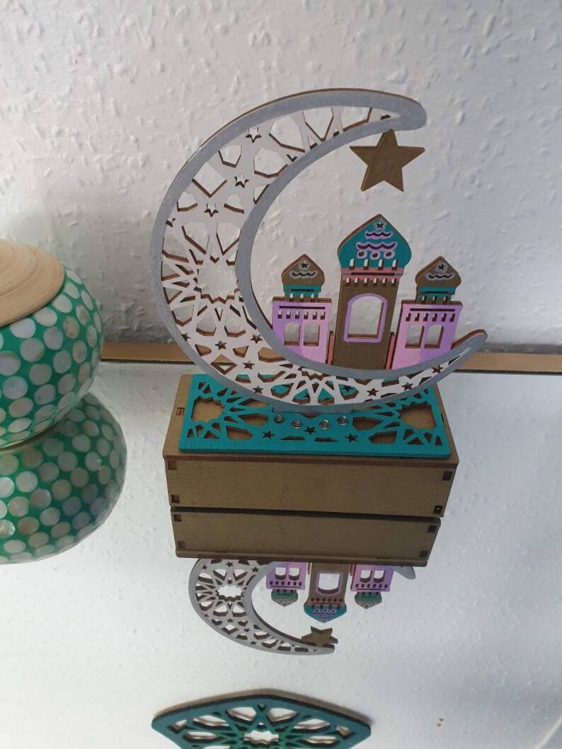 May include: A wooden crescent moon with a star and a small mosque-like structure inside. The moon is decorated with a white and silver geometric pattern. The mosque is painted in pink, teal, and brown. The piece is sitting on a wooden base.