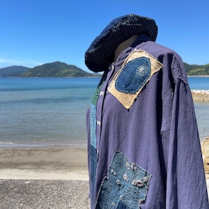 Puede incluir: Una camisa morada de manga larga con varios parches de tela azul y beige que presentan detalles de bordado cosidos a mano. La camisa se combina con una boina azul oscuro que tiene un patrón de puntos blancos en la parte inferior.
