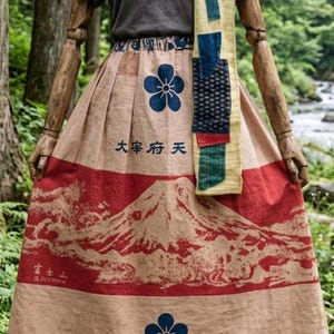 以下が含まれることがあります： グレーのTシャツ、青と緑のパッチが付いた長いベージュのスカーフ、そして赤い山のグラフィック、青い花のモチーフ、"大宰府天"や"富士山"といった日本語の文字がプリントされたベージュのスカート。スカートはウエストがギャザー仕様になっています。
