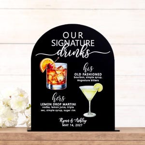 Personalized UV printed acrylic wedding signature drinks sign in black arch shape with white mixed script and block lettering, watercolor cocktail illustrations for his Old Fashioned and hers Lemon Drop Martini with ingredients, couple names and wedding date, displayed on wood bar surface with white florals and bar tools
