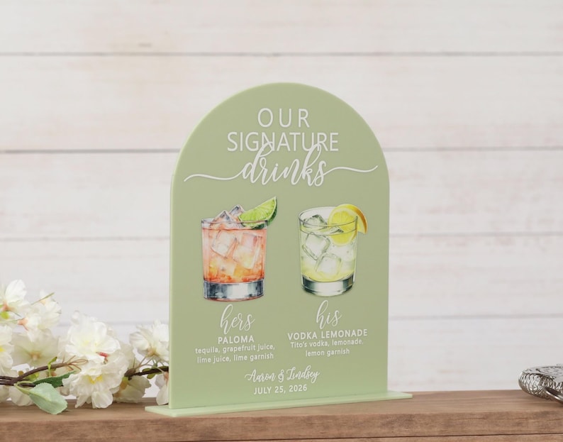 Sage green UV printed acrylic arch wedding signature drinks sign with white lettering and watercolor cocktail illustrations for hers Paloma and his Vodka Lemonade with ingredients listed, personalized with couple names Aaron and Lindsey and wedding date, on wood bar surface with white cherry blossoms and jigger