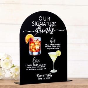 May include: An arched black sign featuring the white text OUR SIGNATURE drinks, his OLD FASHIONED with ingredients, hers LEMON DROP MARTINI with ingredients, and the names Ryan & Ashley with the date MAY 14, 2027, illustrated with two cocktails.