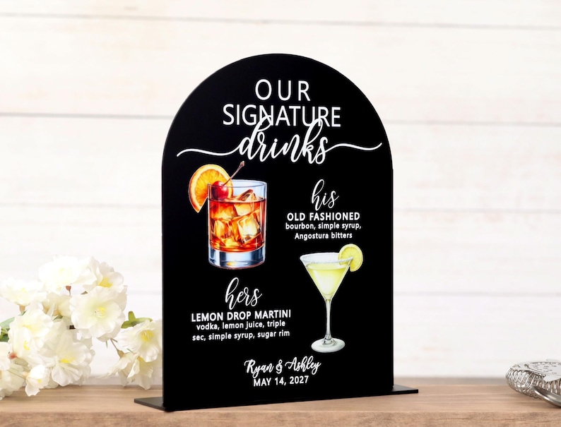 May include: An arched black sign featuring the white text OUR SIGNATURE drinks, his OLD FASHIONED with ingredients, hers LEMON DROP MARTINI with ingredients, and the names Ryan & Ashley with the date MAY 14, 2027, illustrated with two cocktails.