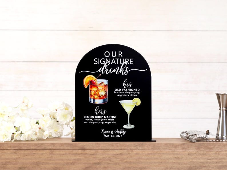 Personalized UV printed acrylic wedding signature drinks sign in black arch shape with white mixed script and block lettering, watercolor cocktail illustrations for his Old Fashioned and hers Lemon Drop Martini with ingredients, couple names and wedding date, displayed on wood bar surface with white florals and bar tools