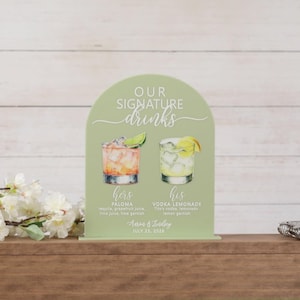 May include: An arched sage green sign featuring the text "OUR SIGNATURE drinks" with watercolor illustrations of a Paloma and a Vodka Lemonade. Below the drinks, the text reads "hers PALOMA", "his VODKA LEMONADE", "Aaron & Lindsey", and "JULY 25, 2026".