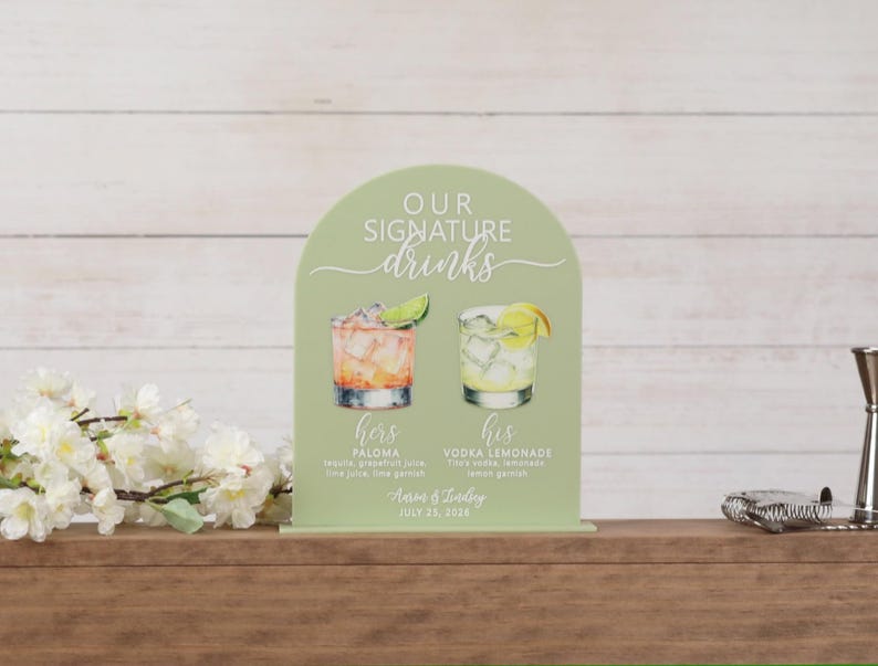 May include: An arched sage green sign featuring the text "OUR SIGNATURE drinks" with watercolor illustrations of a Paloma and a Vodka Lemonade. Below the drinks, the text reads "hers PALOMA", "his VODKA LEMONADE", "Aaron & Lindsey", and "JULY 25, 2026".