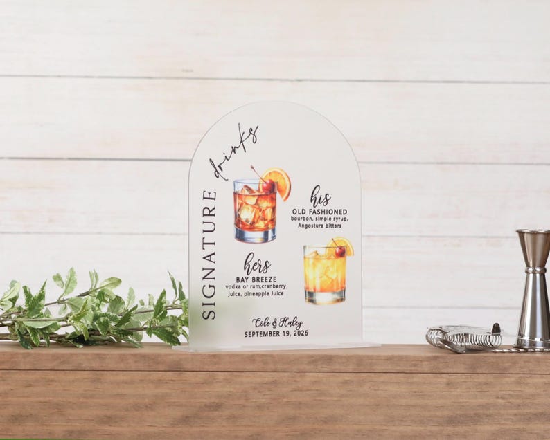 Clear frosted acrylic arch wedding signature drinks sign with vertical script Signature text and watercolor cocktail illustrations for his Old Fashioned and hers Bay Breeze with ingredients, personalized with couple names Cole and Haley and September wedding date on wood bar surface with greenery and bar tools
