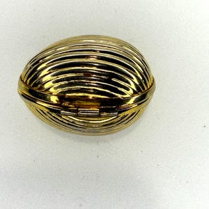 May include: A gold-colored, oval-shaped trinket box with a hinged lid. The box features a ribbed design, with horizontal lines creating a textured surface. The box is closed and sits on a white surface.