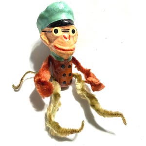 May include: A vintage monkey toy with a green hat, black rim, and round glasses. The monkey has an orange body, arms, and legs. The legs are made of fuzzy, light-yellow material. The toy has a painted face with a red mouth.