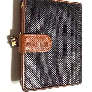 May include: A small, rectangular wallet with a textured black surface and brown leather accents. The wallet has a brown leather clasp and gold-colored metal hardware. The edges are trimmed with brown leather.