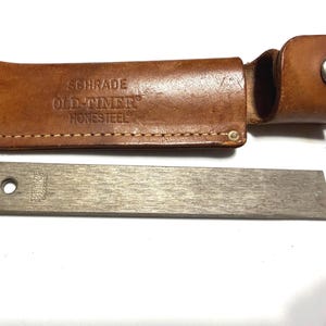 May include: A brown leather sheath with the text "SCHRADE OLD-TIMER HONESTEEL" embossed on the front. A silver-colored metal sharpening steel is next to the sheath. The sharpening steel has two holes near the top and the text "SCHRADE HONESTEEL" printed on it.
