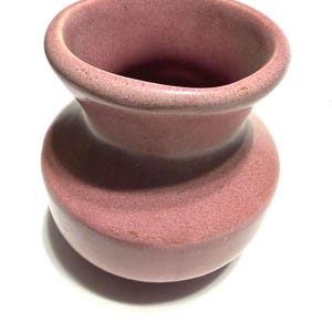 May include: A dusty rose-colored ceramic vase with a rounded body and a flared, open top. The vase has a textured surface and a classic, hourglass shape. The interior is a darker shade of the same rose color.