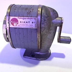 May include: Vintage gray pencil sharpener with a hand crank. The sharpener has a rectangular body with a label that reads "Apseco GIANT 51" and "Beverly Hills, Calif. USA."