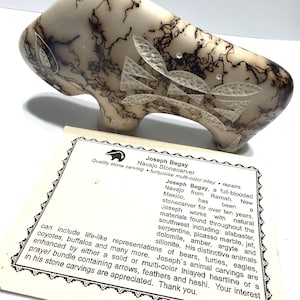 May include: A carved stone buffalo sculpture with etched details and clear, leaf-shaped accents. The sculpture is off-white with black veining. A card with text about the artist, Joseph Begay, is in the background.