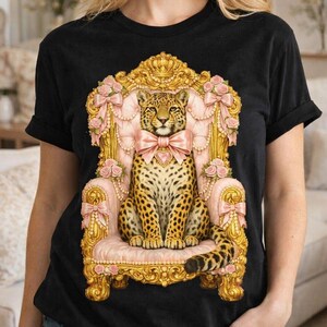 Puede incluir: Camiseta negra con un leopardo con pajarita rosa, sentado en una silla adornada rosa y dorada. La silla está decorada con lazos y rosas rosas. El leopardo tiene un pelaje manchado y una larga cola.