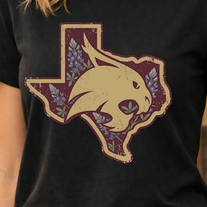 Puede incluir: Camiseta negra con un gráfico desgastado del estado de Texas en granate, con un contorno beige y una cabeza de gato montés beige. El gráfico incluye detalles florales azules y morados, creando un aspecto vintage.