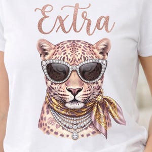 May include: White t-shirt with a leopard illustration wearing sunglasses, a scarf, and jewellery. The word "Extra" is written above the leopard in a rose gold glitter font. The leopard has a spotted coat.