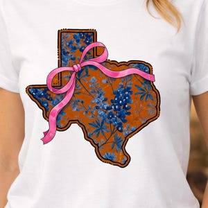 May include: White t-shirt featuring a Texas-shaped design with a floral pattern in shades of blue and orange. A pink ribbon is tied across the design. The shirt is a casual wear item.