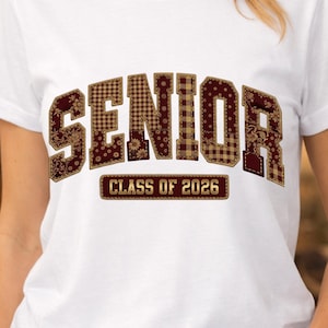 May include: White t-shirt featuring the word "SENIOR" in a patchwork design with burgundy and gold accents. Below the word "SENIOR" is the text "CLASS OF 2026" in a rectangular box. The shirt is a classic crew neck.
