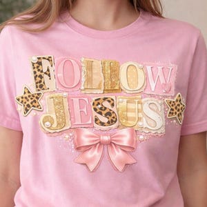 Puede incluir: Camiseta rosa con la frase "FOLLOW JESUS" en un diseño decorativo. El texto incorpora estampado de leopardo, purpurina dorada y un lazo rosa. Estrellas con detalles de estampado de leopardo flanquean el texto.