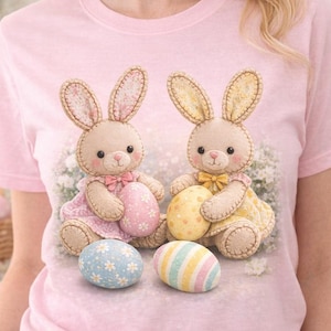 Puede incluir: Camiseta rosa claro con un diseño de dos conejitos de Pascua de peluche sosteniendo huevos decorados. Los conejitos llevan vestidos y lazos. Dos huevos de Pascua decorativos están frente a los conejitos.