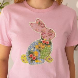 Puede incluir: Camiseta rosa con un diseño de conejo de patchwork. El conejo tiene orejas con estampado floral, un cuerpo floral azul y amarillo y una sección de lunares verdes. El diseño está delineado con un borde de purpurina plateada.
