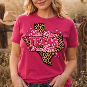 Puede incluir: Una camiseta magenta con un contorno de Texas con estampado de leopardo y el texto "She's From TEXAS I Can Tell" en rosa y dorado. La camiseta de manga corta tiene cuello redondo. El diseño está centrado.