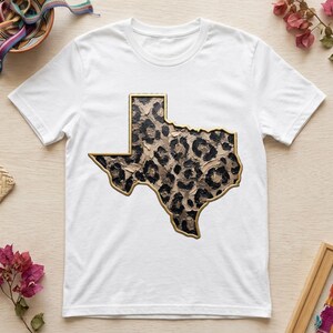 Puede incluir: Una camiseta blanca de manga corta con un gráfico central del estado de Texas. La forma del estado está rellena con un estampado de leopardo texturizado en negro y beige, y está delineada con un borde dorado metálico.