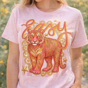 Puede incluir: Camiseta rosa claro con un diseño de tigre y la palabra "Easy" en naranja. El tigre presenta tonos rosas y naranjas, sobre un fondo decorativo dorado y rosa con estrellas. Una camiseta casual para el día a día.